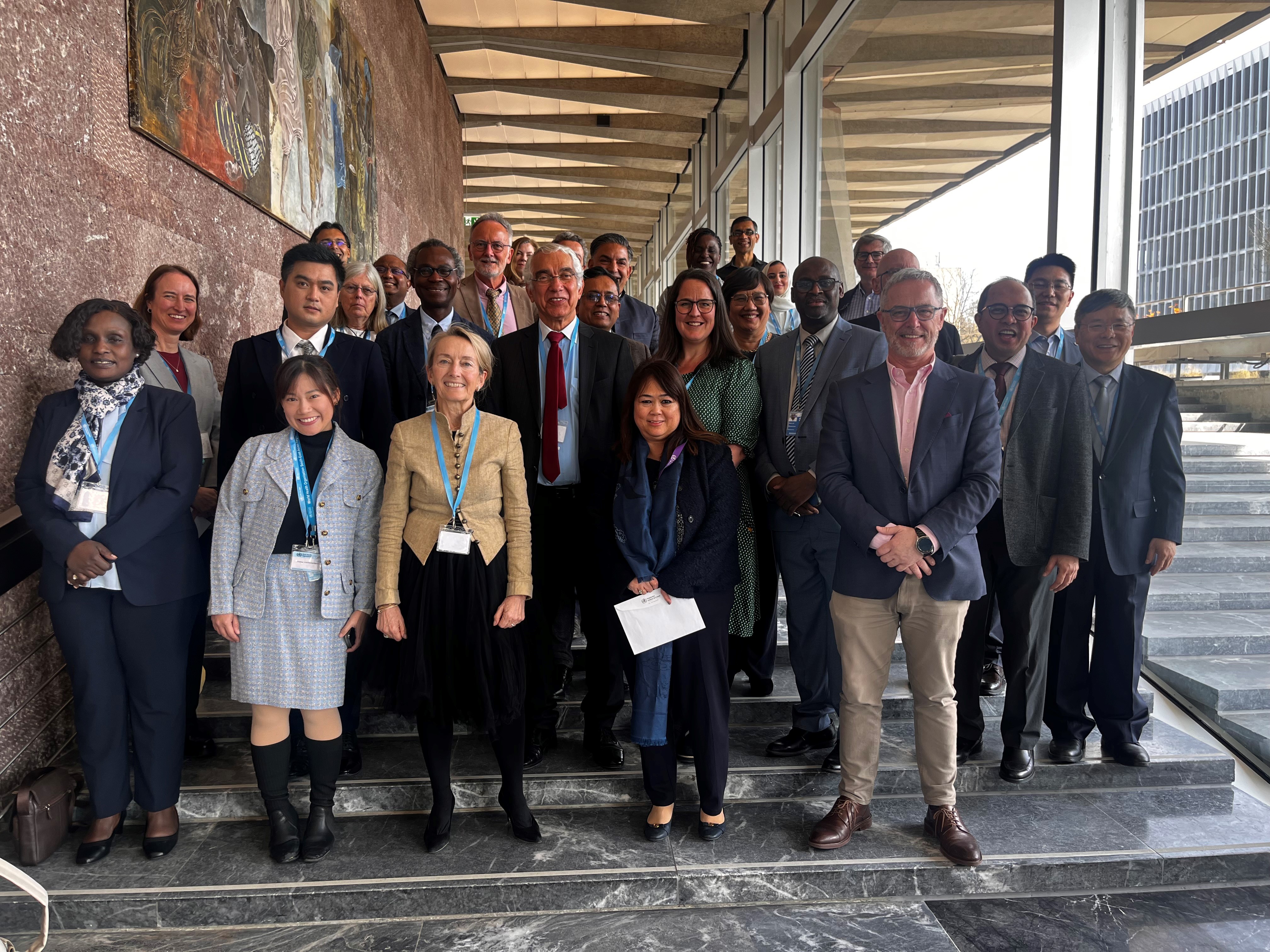 Group of WHO steering committee members in business attire posing on steps in a modern building corridor.