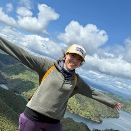 Photo of Javiera Perez smiling and standing with her arms wide open as she stands on a cliff overlooking water