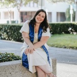 Photo of Jennifer Riekes sitting with her legs crossed on a planter outside at UC Irvine. She is wearing a blue and yellow graduation sash.