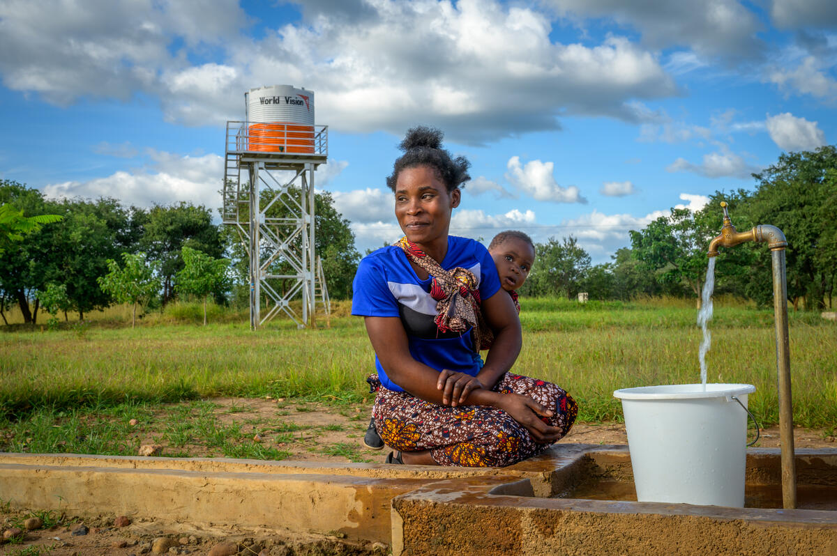 Woman sitting with her baby outside by a field and a spicket of water that is filling up a bucket