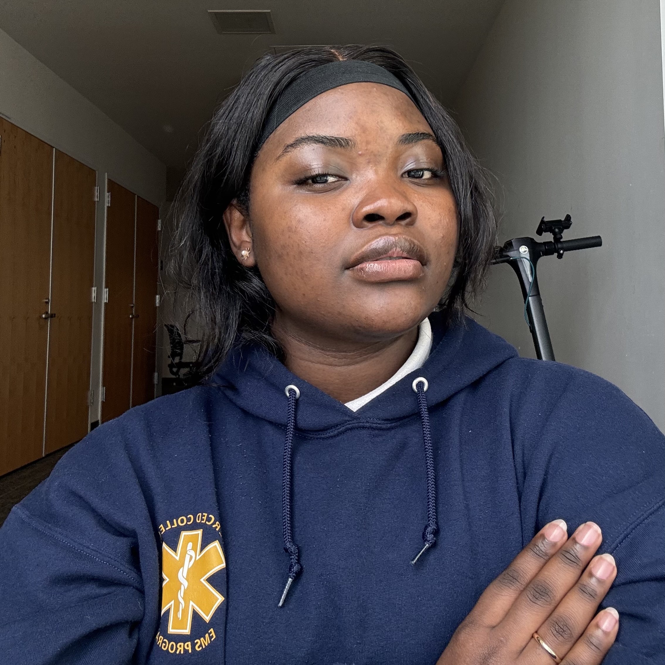 A young woman, wearing a navy hoodie with a logo, poses with her arms crossed in a indoor setting.