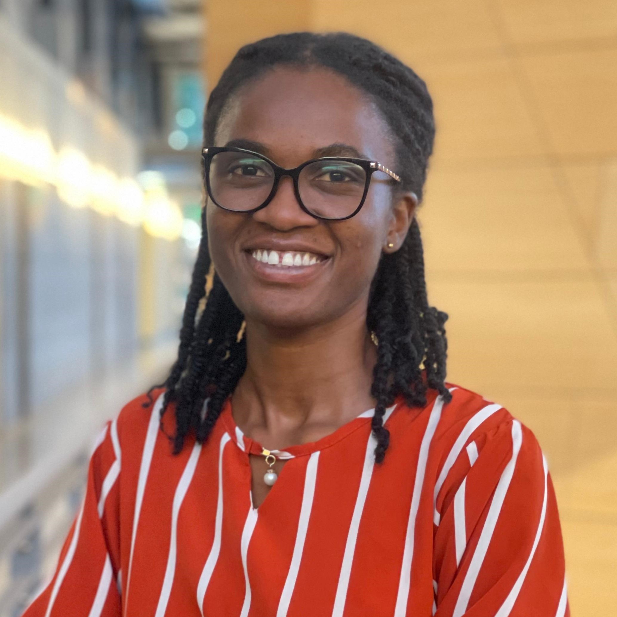 A smiling young woman with glasses wearing a red and white striped blouse, standing in a modern indoor setting.