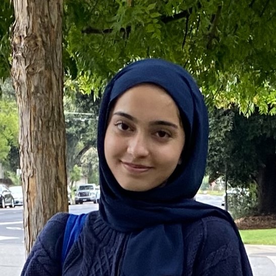 A smiling young woman wearing a navy blue hijab and sweater stands outdoors near a tree.