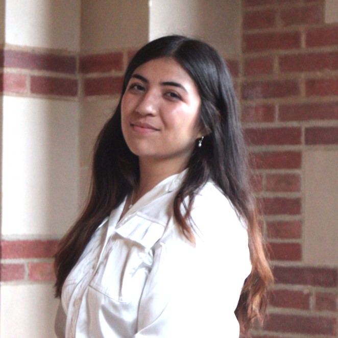 A young woman with long dark hair, smiling and wearing a white jacket, stands in front of a brick wall.