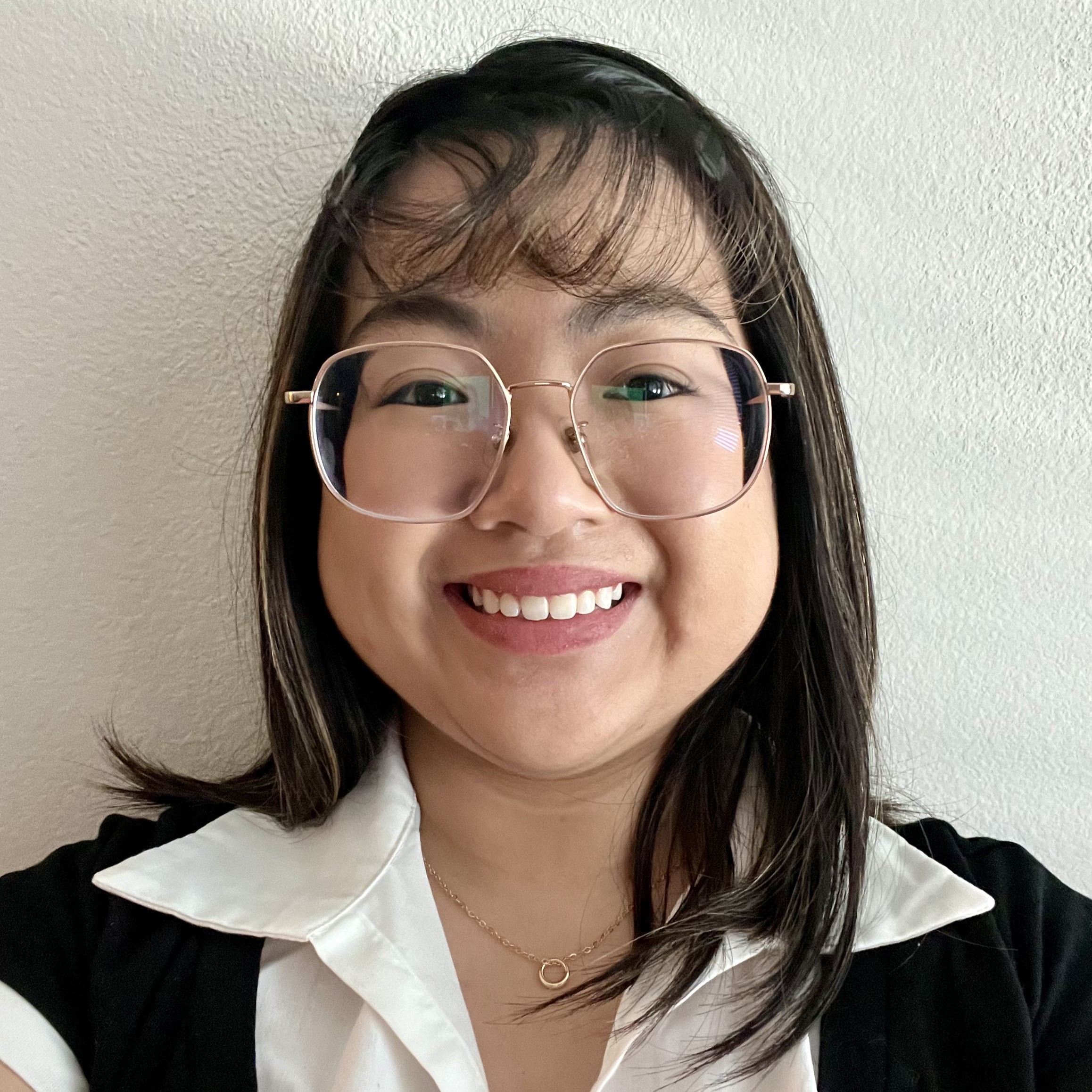A smiling woman with glasses and long hair, wearing a white shirt under a black vest, standing against a light-colored wall.