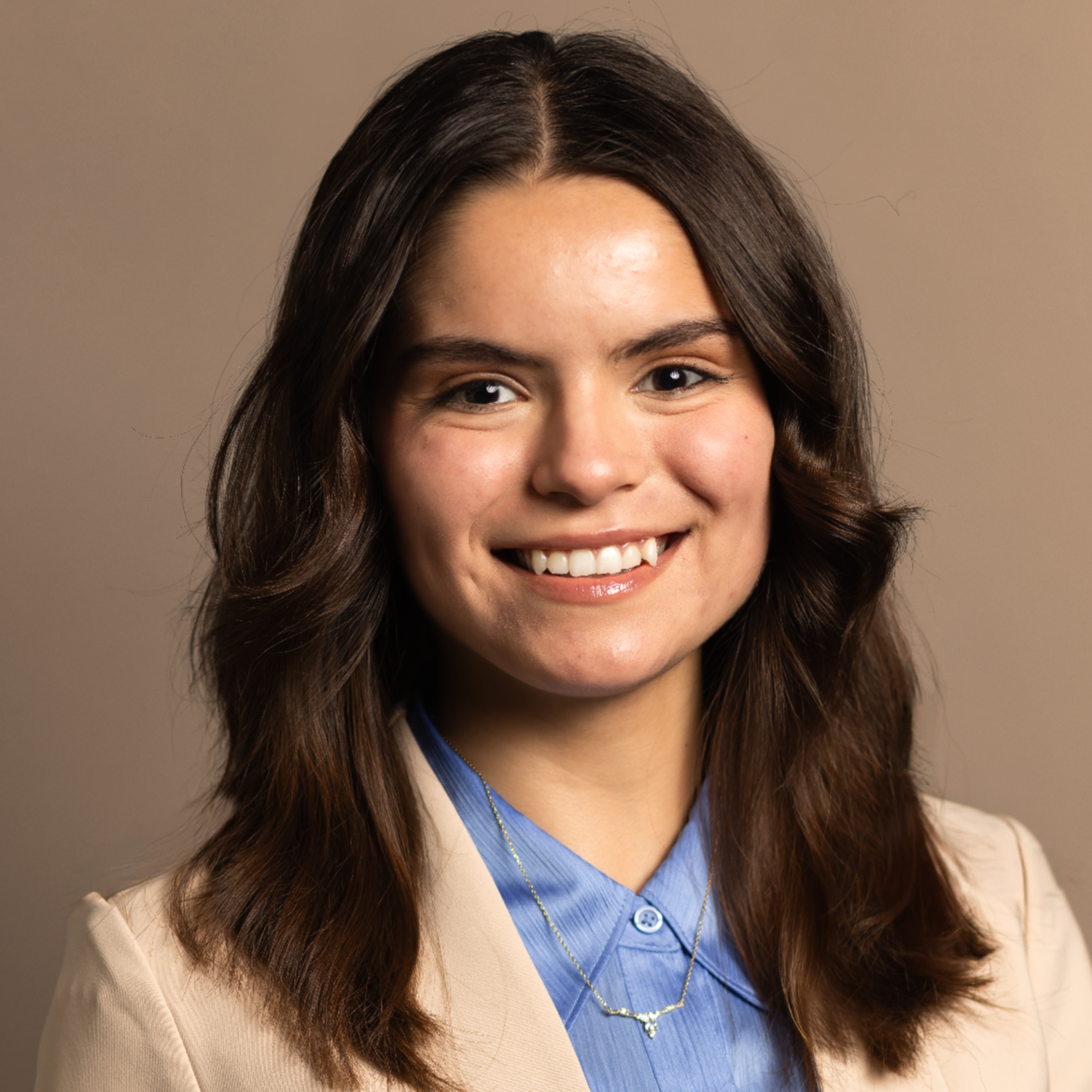A professional portrait of a young woman with long, wavy hair, wearing a light blazer and a blue shirt, smiling confidently.