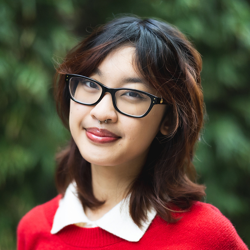 Headshot of a young woman wearing black rimmed glasses and a red sweater with a white collared shirt layered under and standing in front of greenery.