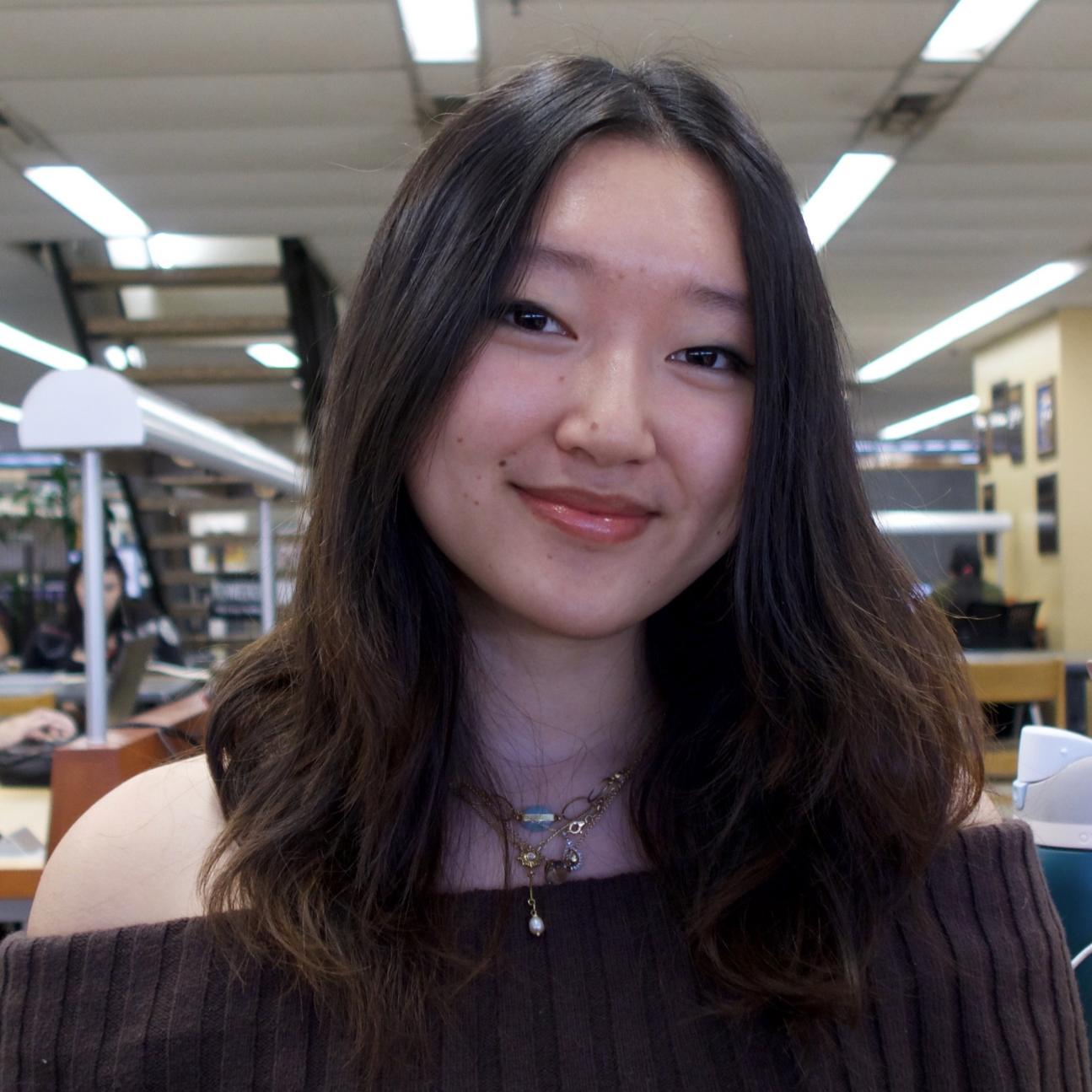 A young woman with long, wavy hair smiles warmly while sitting in a library setting.