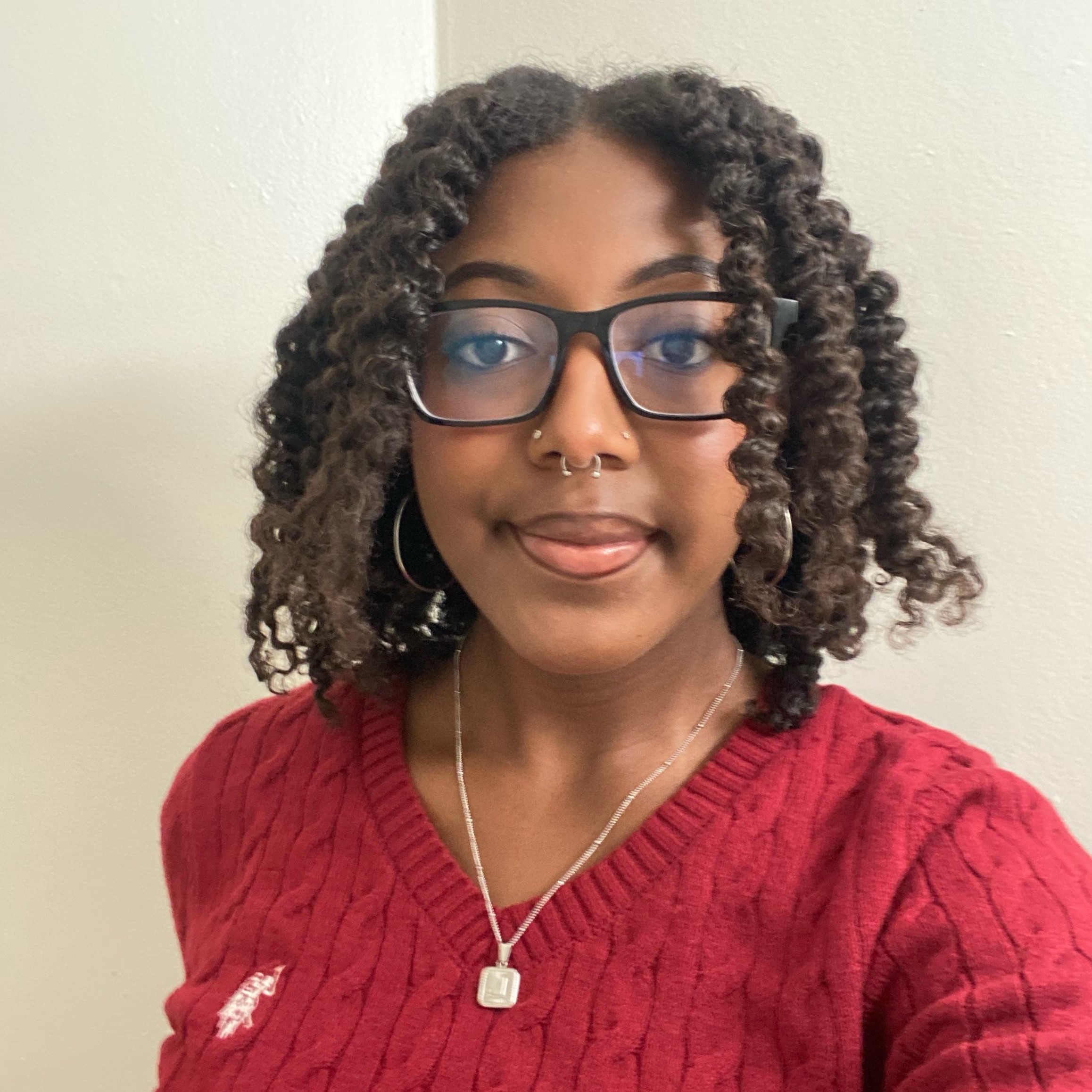 A young woman with curly hair and glasses wearing a red sweater, smiling lightly at the camera.