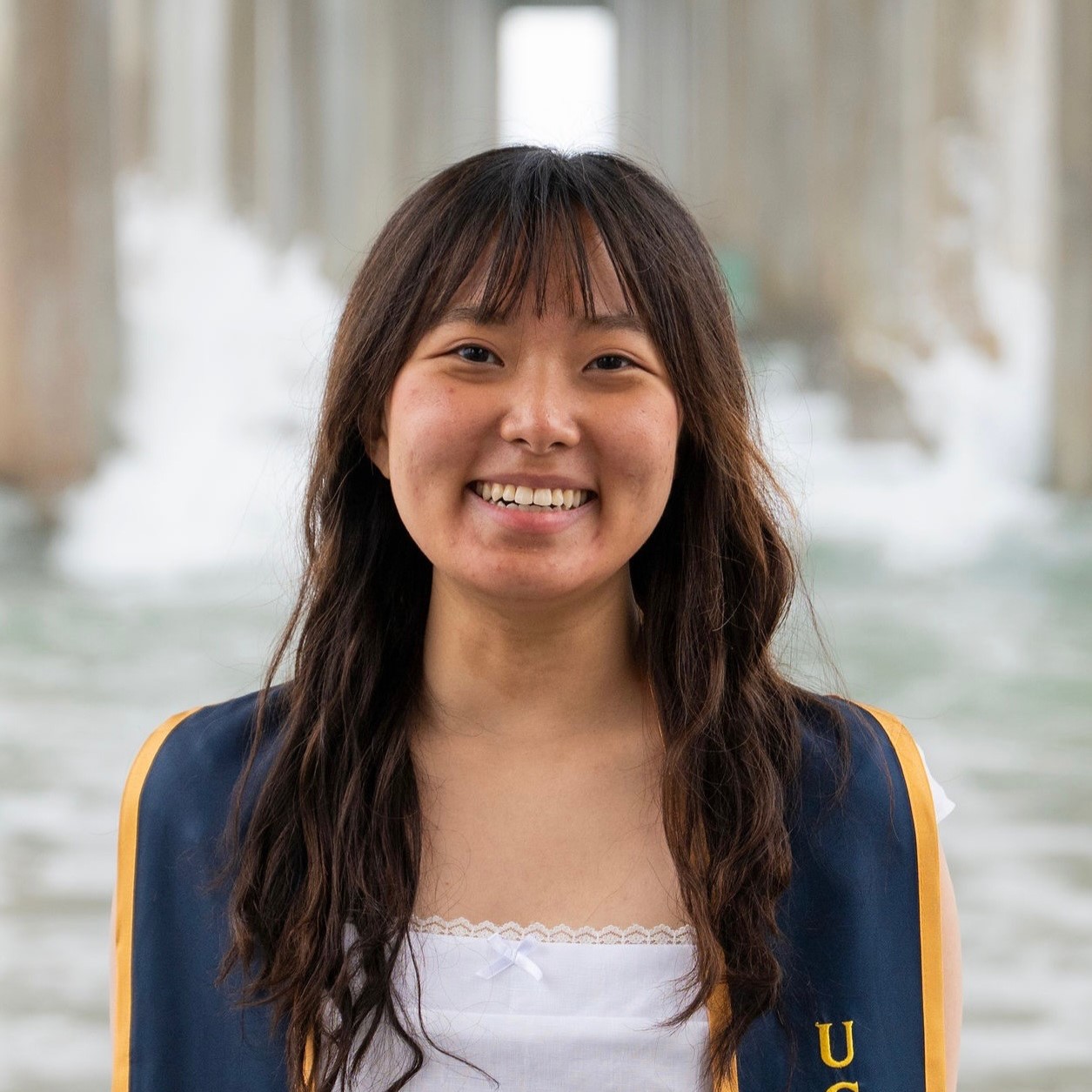 A smiling graduate wearing a blue academic gown stands in front of a pier with waves in the background.