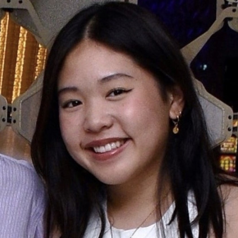 Smiling young woman with long dark hair, wearing a white top and gold earrings, in a vibrant setting.