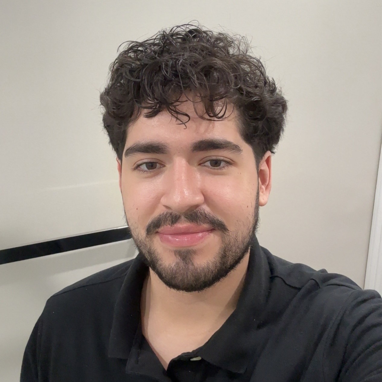 A young man with curly hair and a beard smiles while wearing a black shirt.