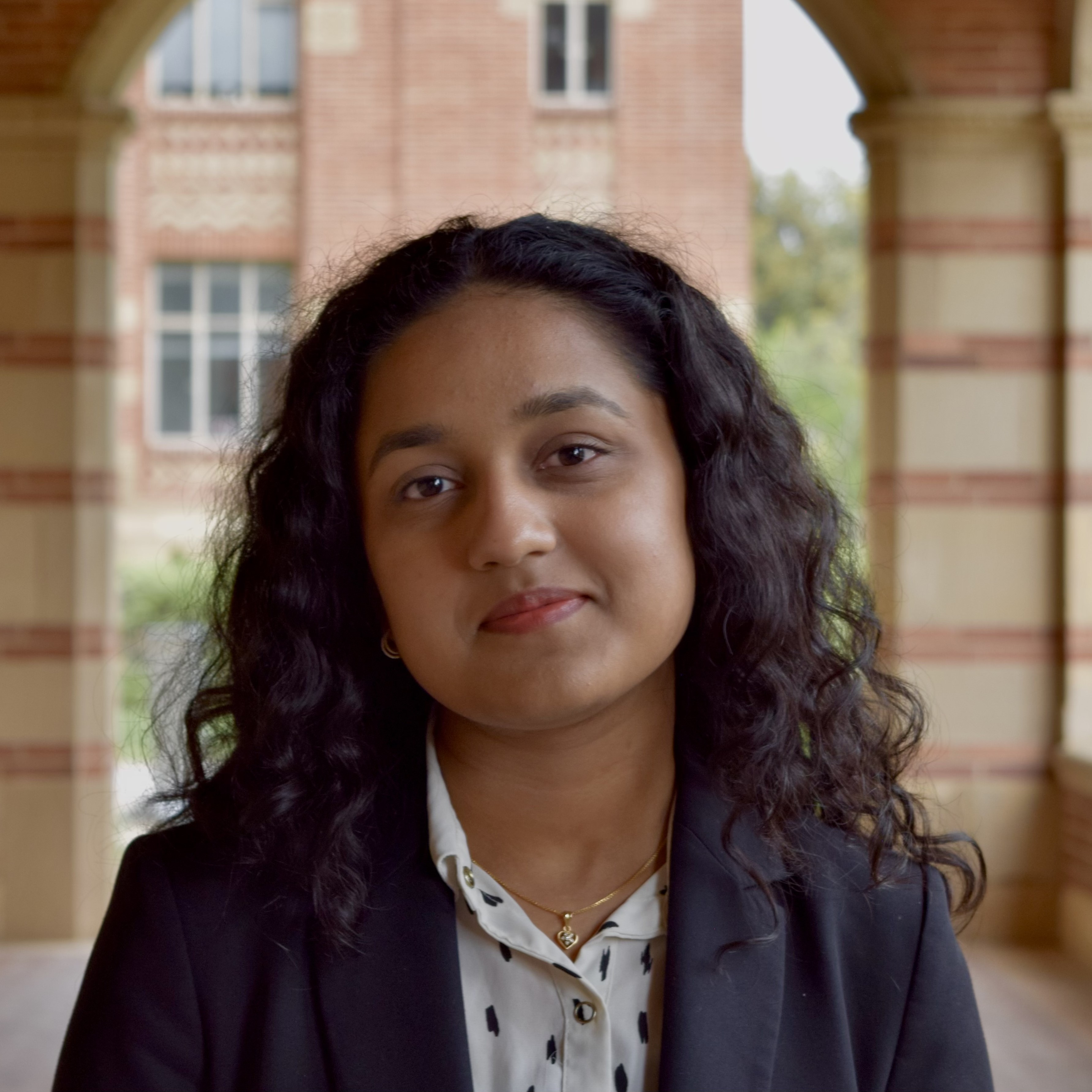 A young woman with curly hair is smiling confidently while wearing a blazer, set against a backdrop of a brick building and archway.