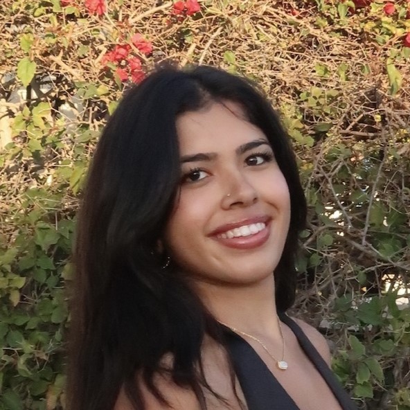 A smiling young woman with long dark hair standing outdoors amidst green foliage and flowering plants.