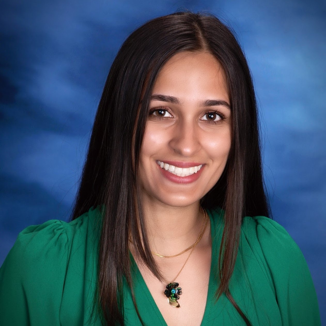 A smiling woman with long black hair wearing a green blouse and a necklace, posed against a blue background.