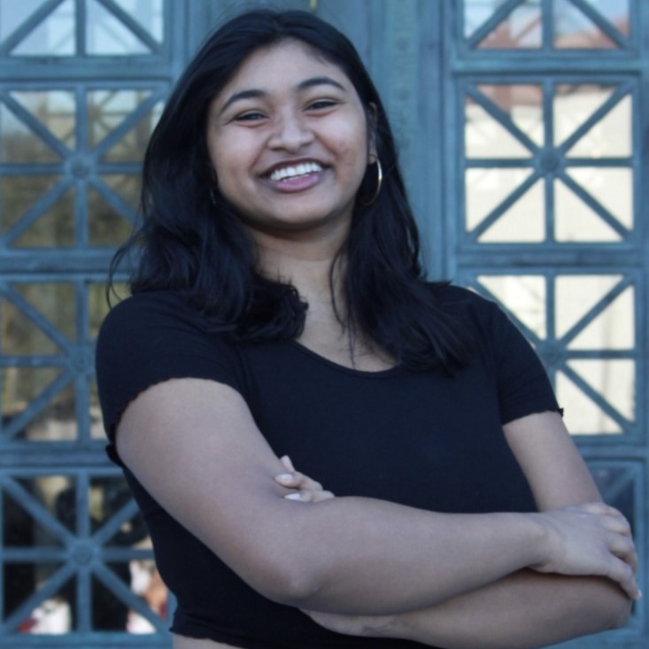 A smiling young woman with long black hair stands confidently with her arms crossed in front of a patterned window.