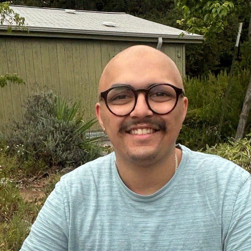 A smiling person with a bald head and glasses sitting outside, surrounded by greenery.