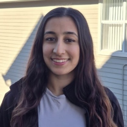 A smiling young woman with long dark hair, wearing a black jacket and a light shirt, standing outside.