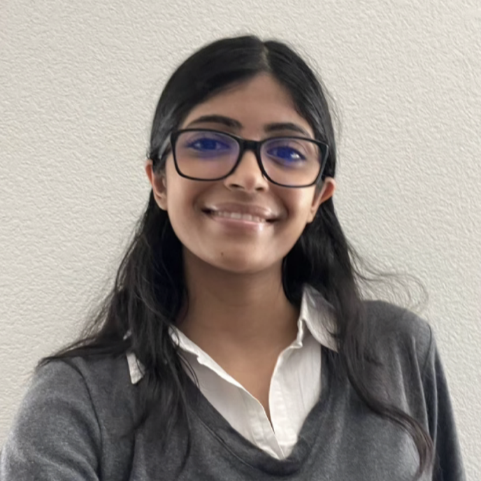 A young woman with long dark hair and glasses, smiling while wearing a gray sweater and white blouse against a plain background.
