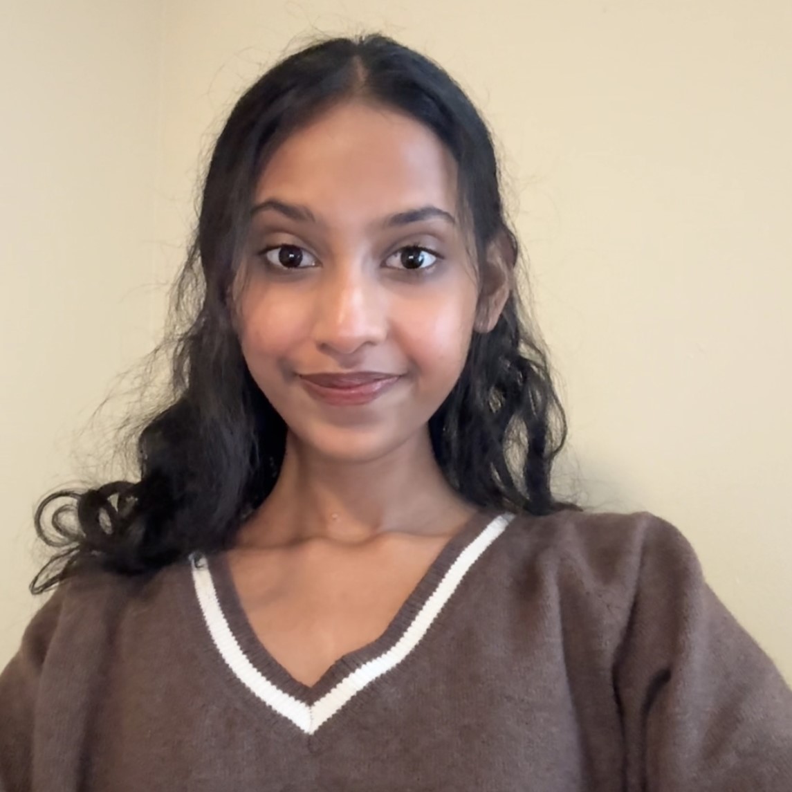 A smiling young woman with long, wavy hair wearing a brown sweater with a white V-neck.
